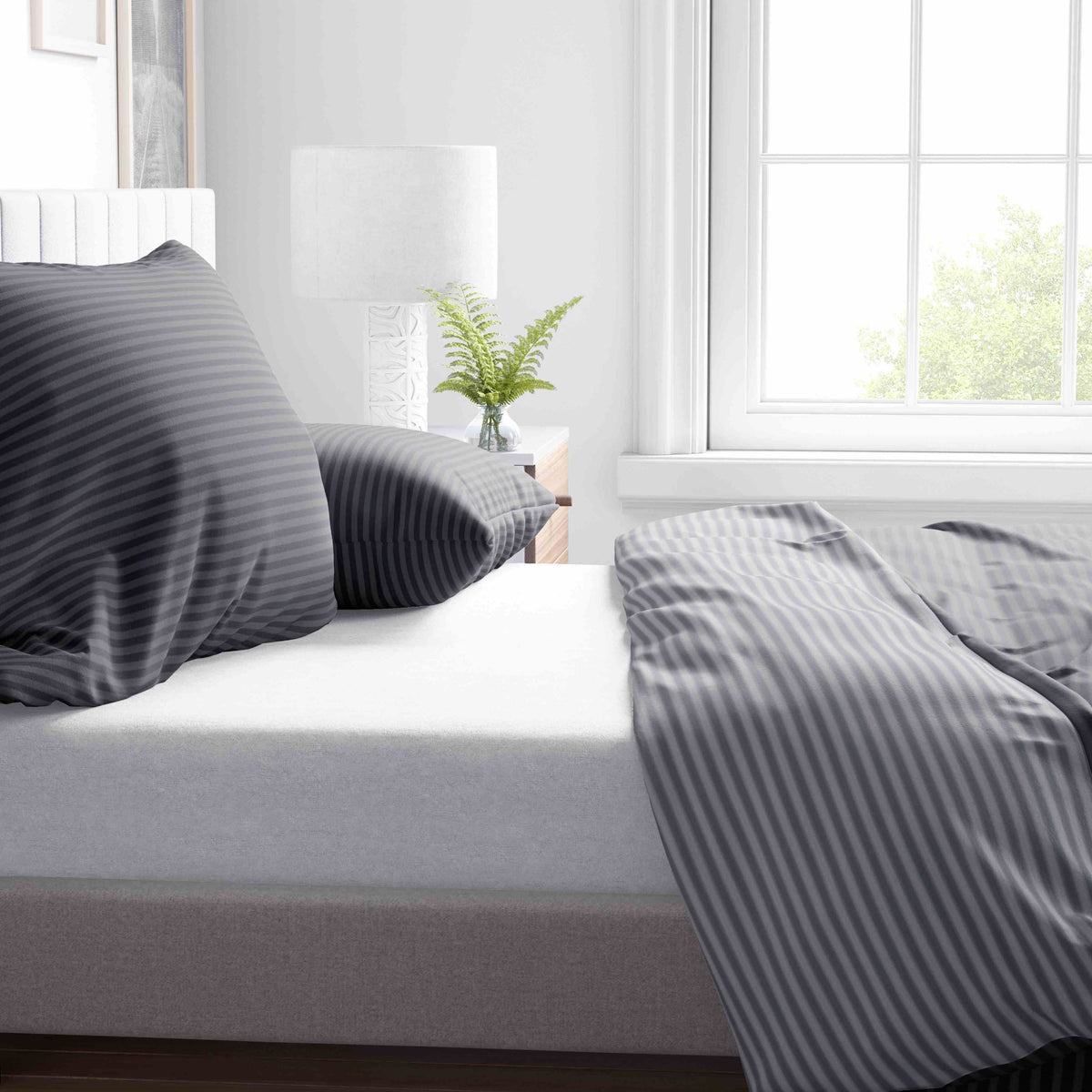 "Nahaufnahme eines Bettes mit Wendebettwäsche aus 100% Baumwolle Renforce in Anthrazit Grau mit Streifen Design, flankiert von einem Nachttisch mit Lampe und einer Pflanze."